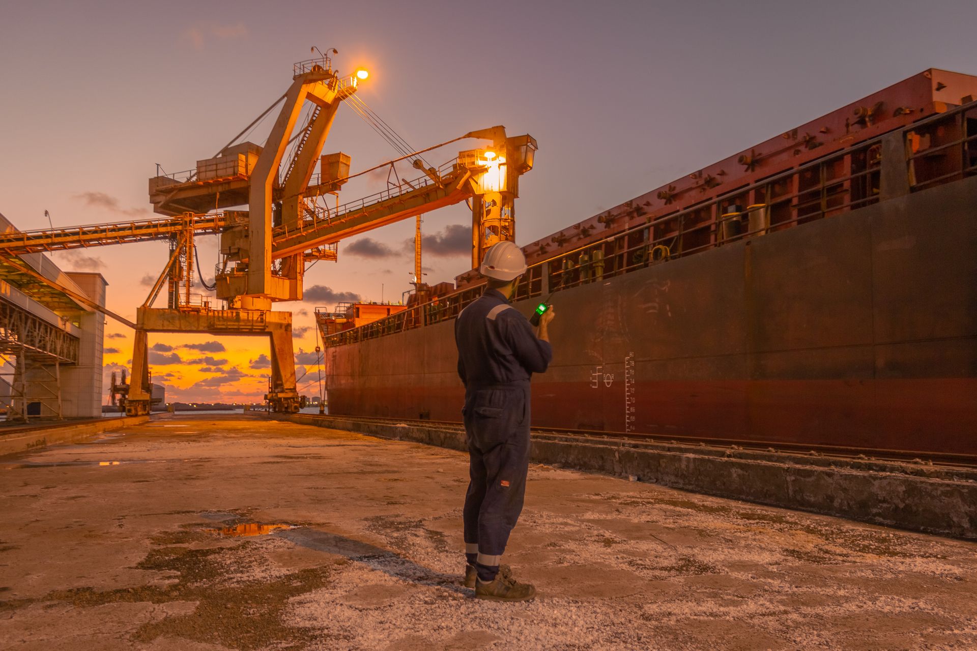 Supervisore portuale con casco che segue il carico di una nave rinfusiera al tramonto